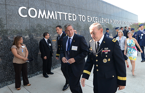 An image of well-dressed persons in an area with letters on a wall behind that read. Committed to excellence in defense of the nation.